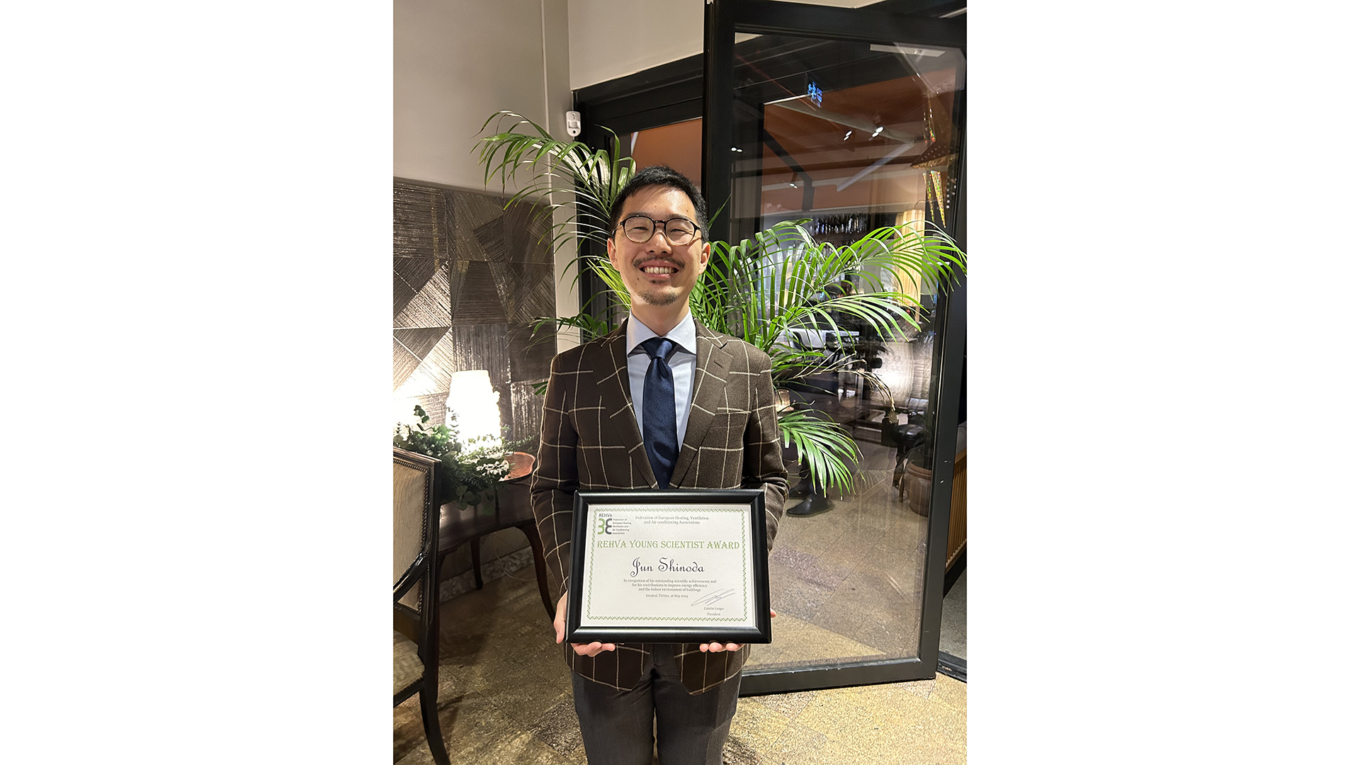 A gentleman wearing a brown suit posing for a photo while holding his diploma